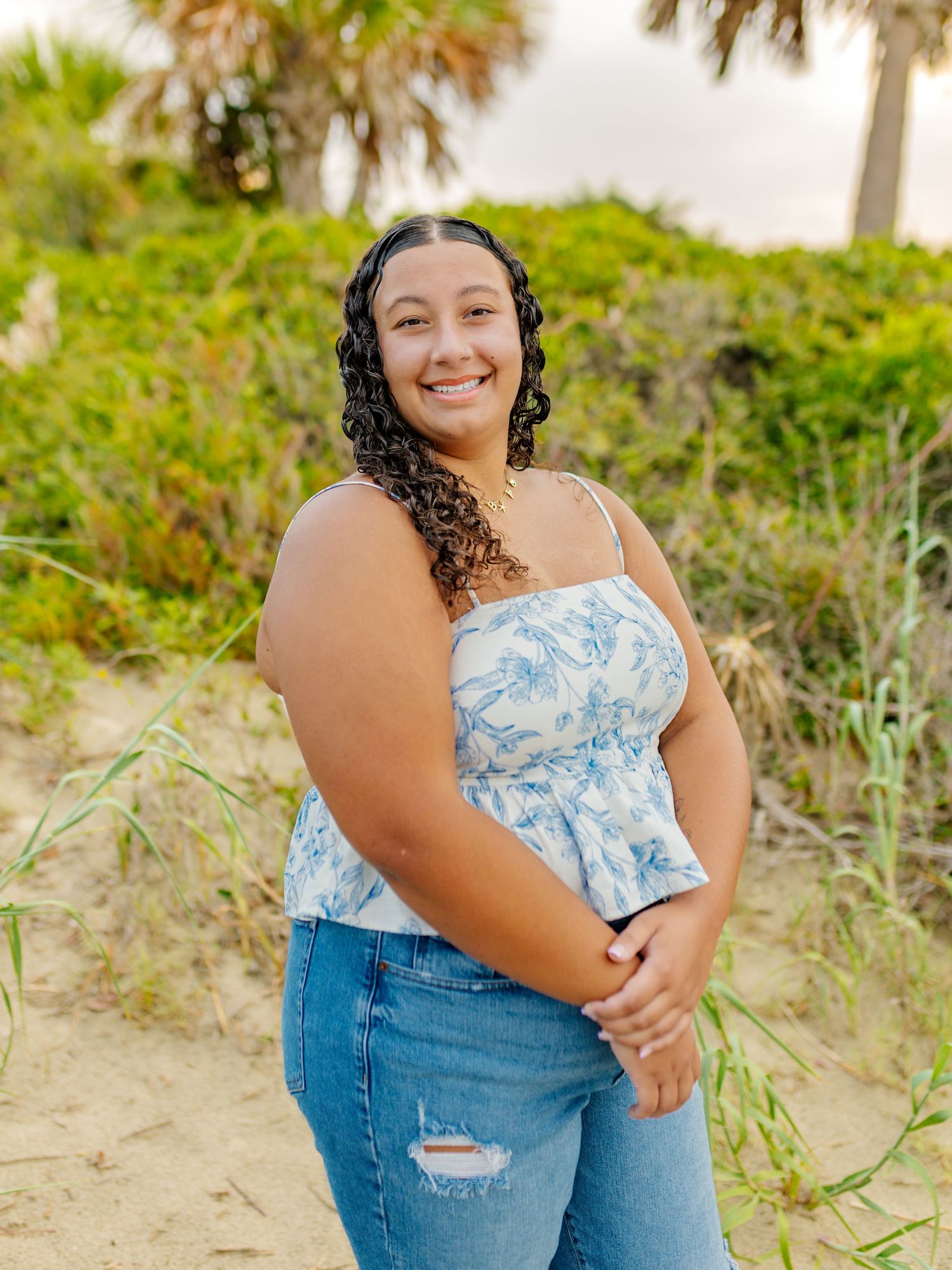Woman in floral top and jeans poses on beach with palm trees in background.