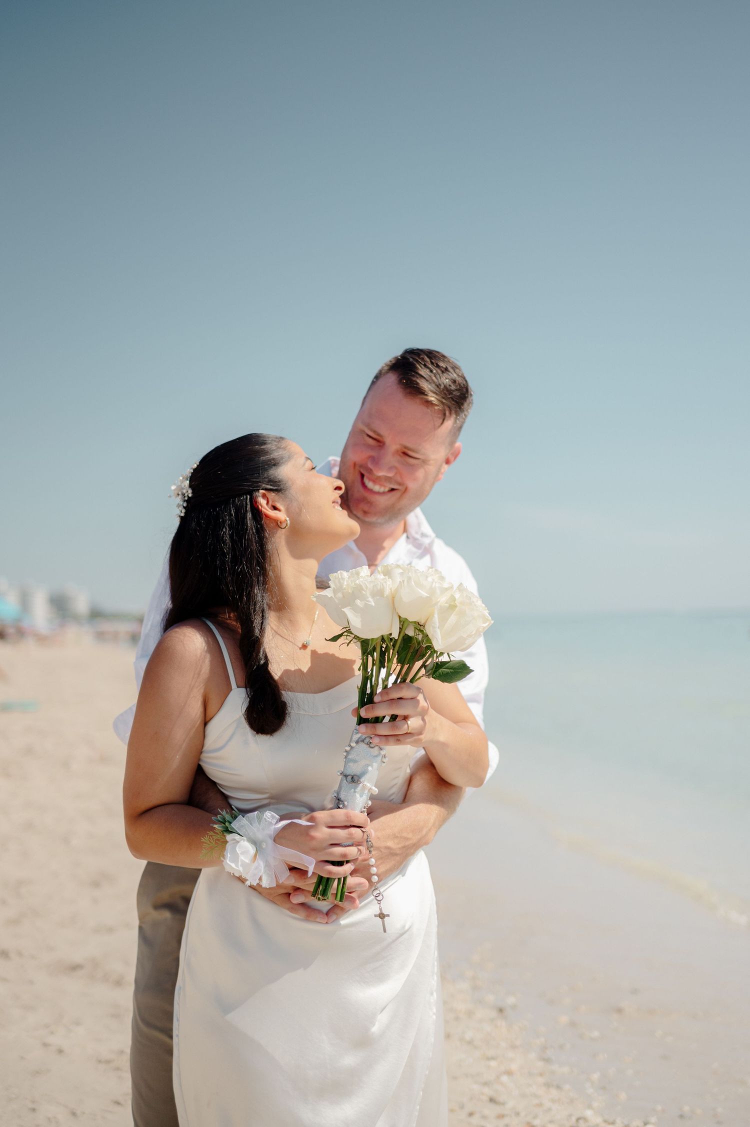 A romantic couple shares tender moments on a sunny beach with white flowers and tropical ocean views in the background.