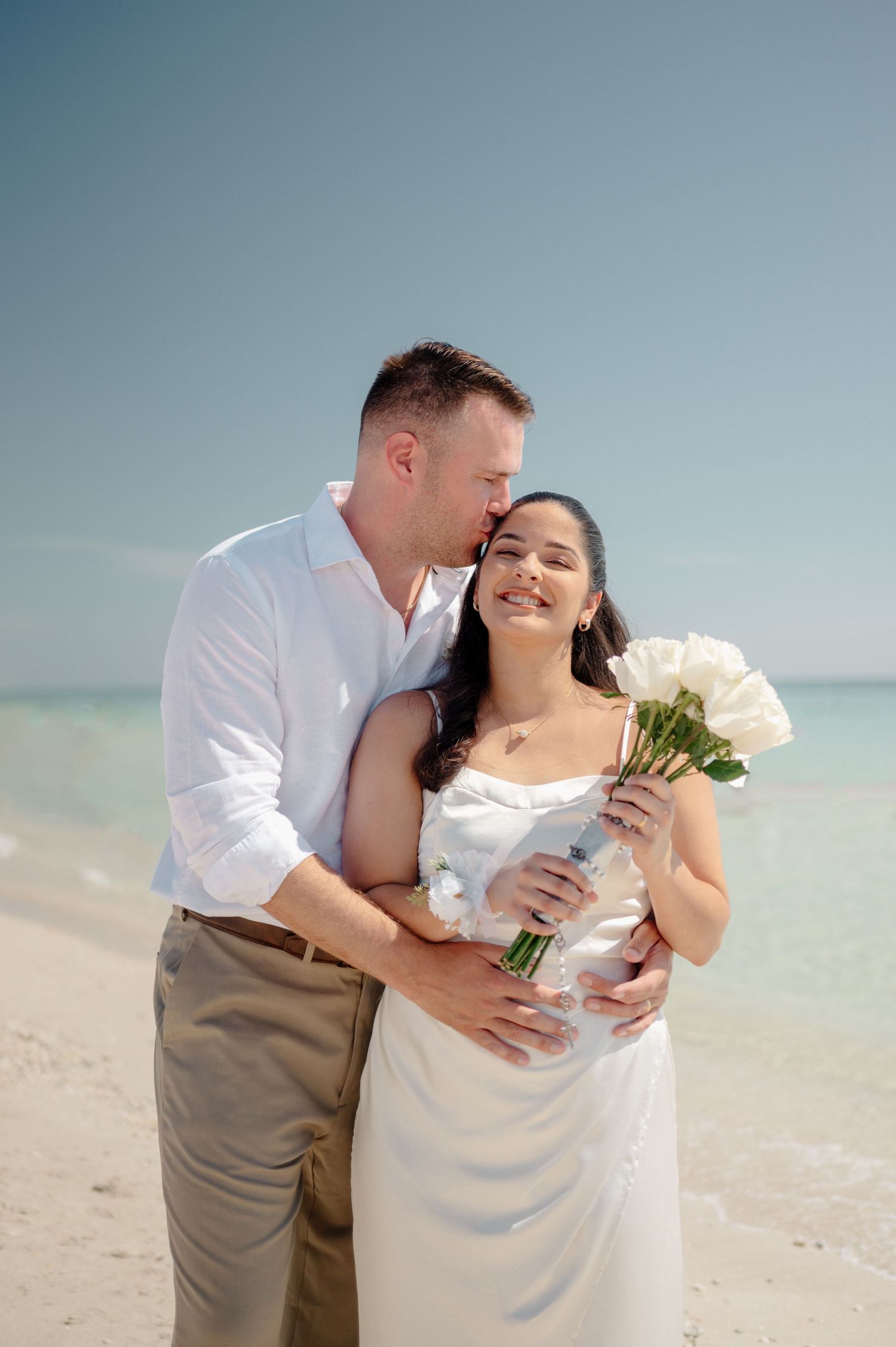 Romantic beach wedding couple embraces while holding white roses against turquoise ocean backdrop.