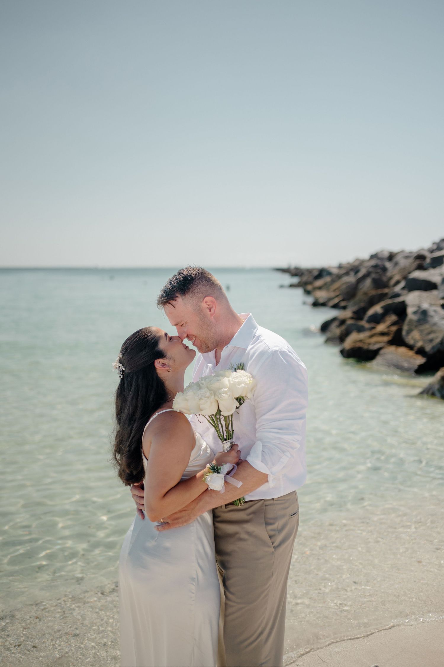 A romantic couple embraces on a rocky beach shoreline with crystal clear waters in the background.
