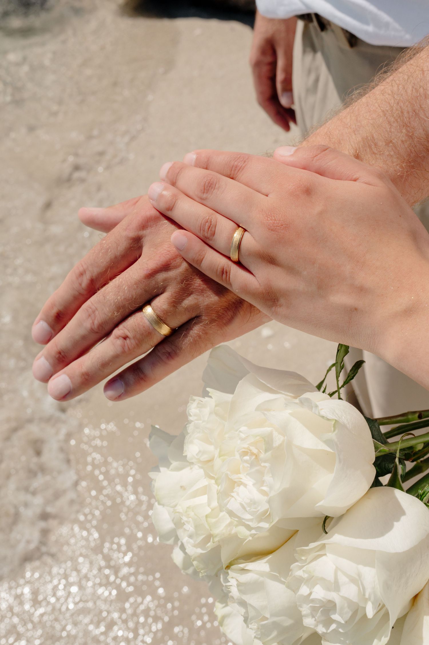 A pair of hands with a wedding ring arranges delicate white peonies on a light surface.