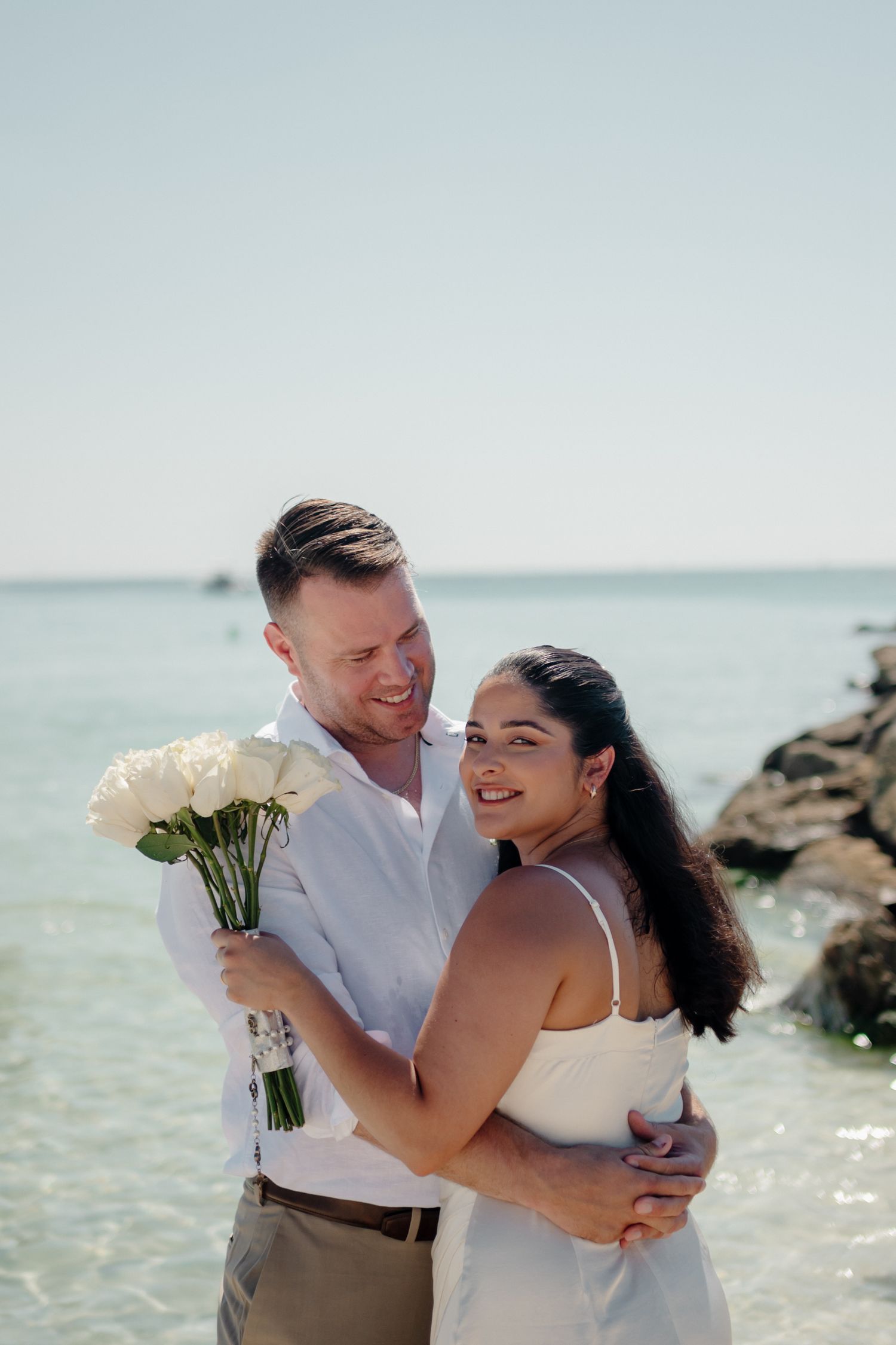 A happy couple embraces on a sunny beach with white roses, wearing light summer wedding attire by the ocean.