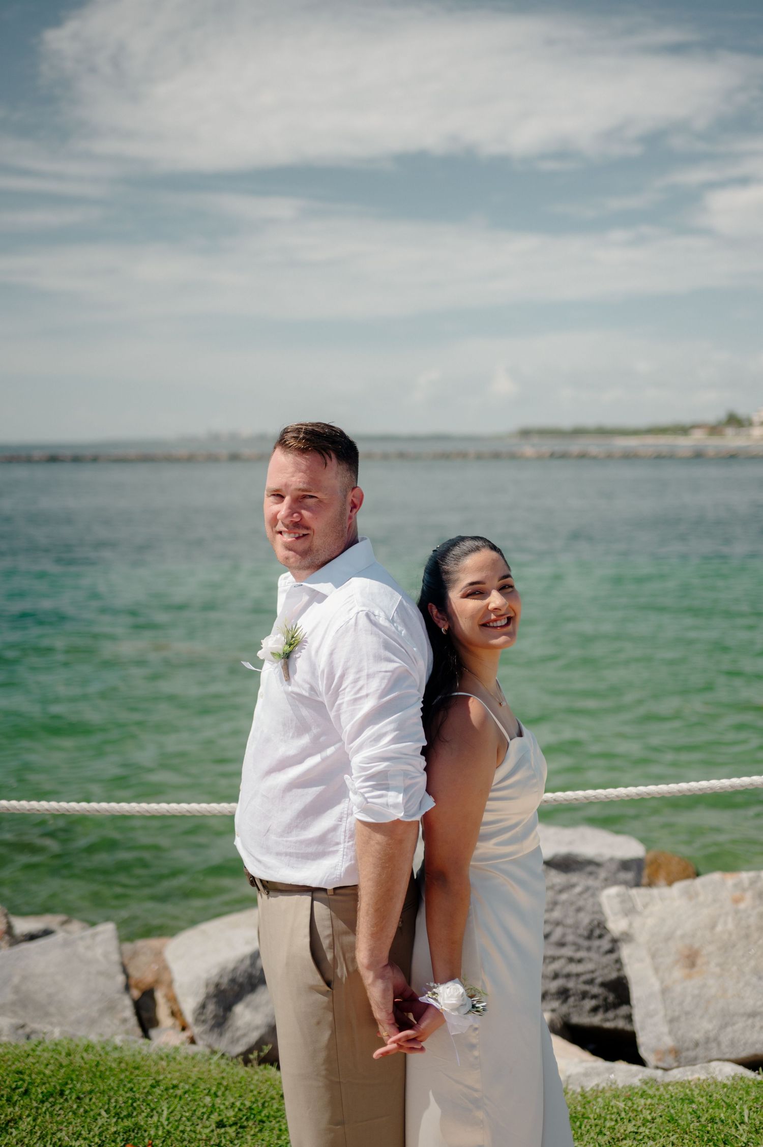 A couple poses together on a rocky shoreline with turquoise ocean waters and cloudy skies in the background.