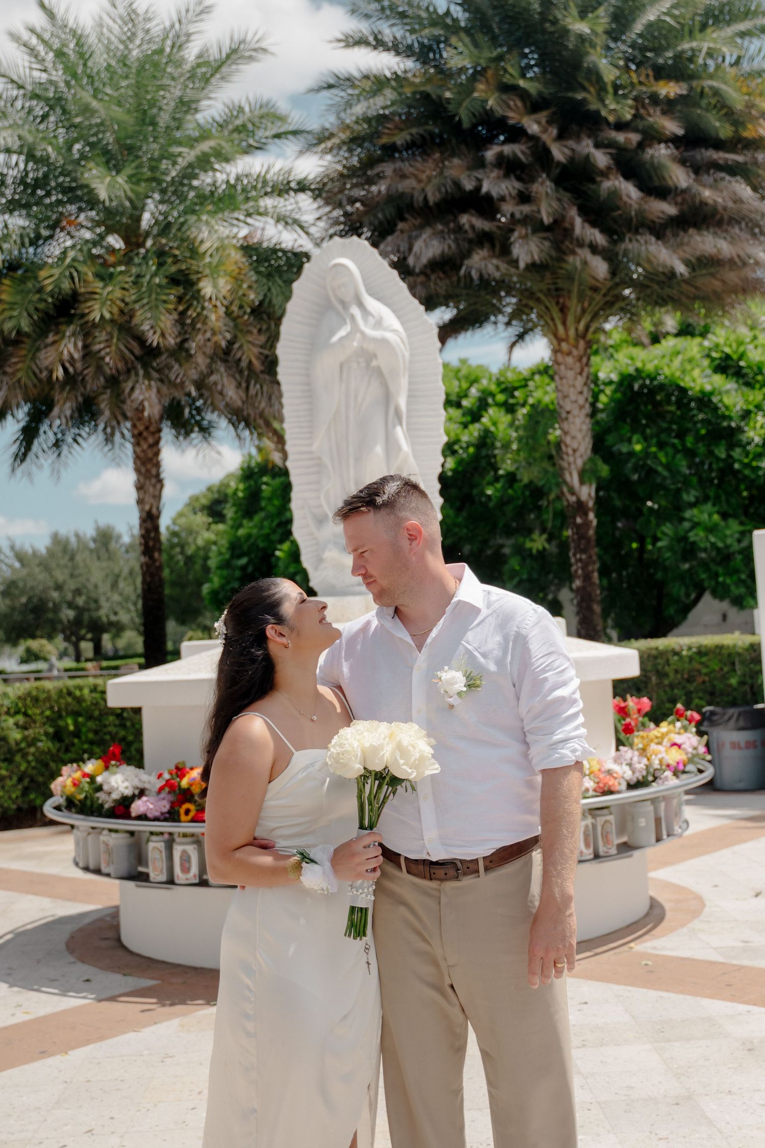 A couple shares a romantic kiss under palm trees at a tropical wedding venue with white decor and flowers.