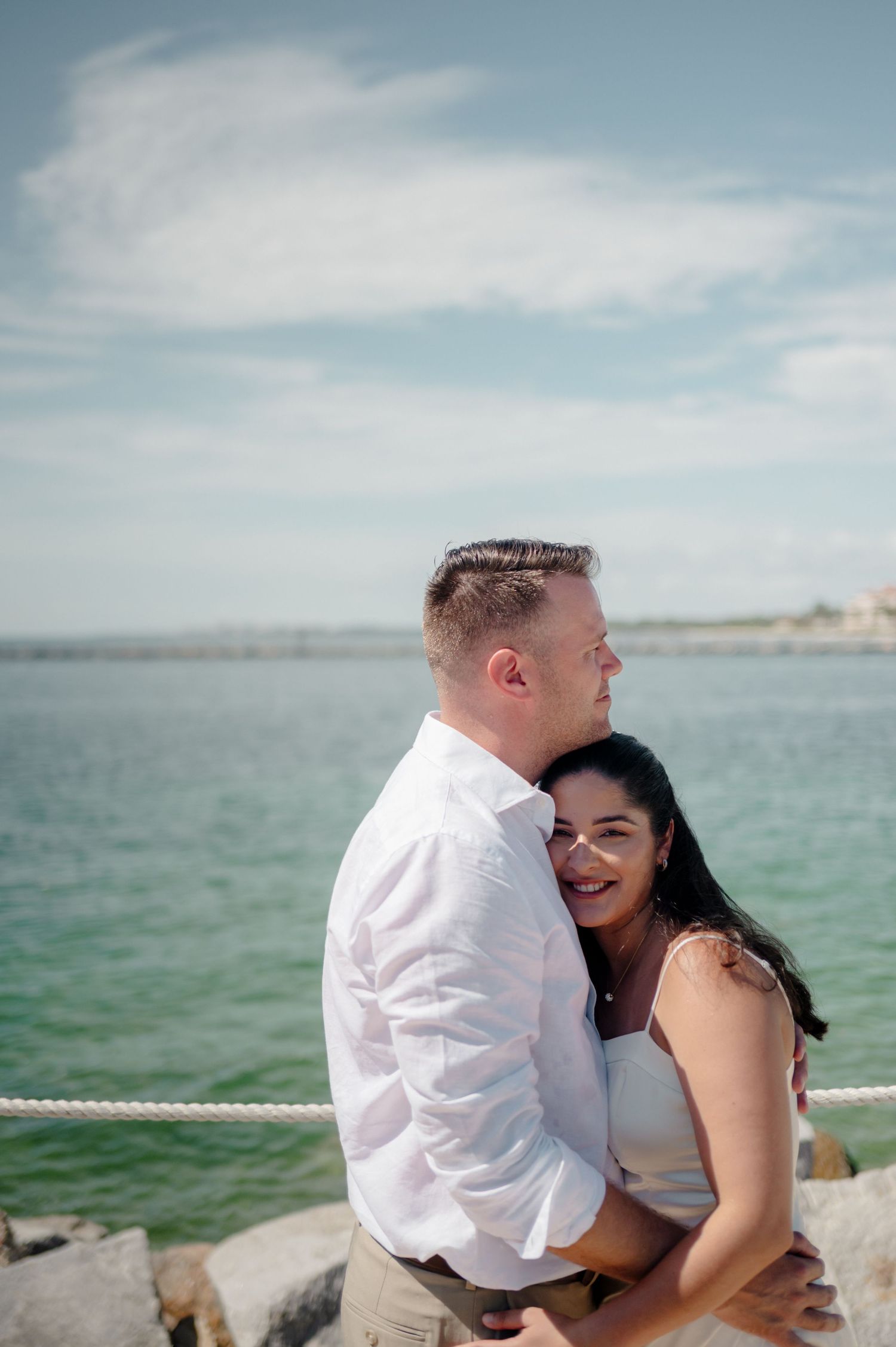 A couple shares romantic moments by the ocean with turquoise waters and cloudy skies in the background.