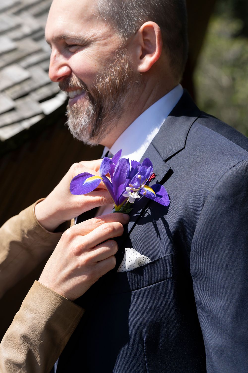 A purple iris boutonniere is being pinned to a formal suit jacket in natural sunlight.