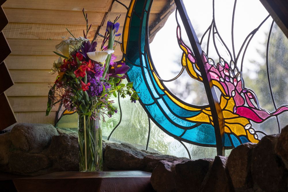Colorful stained glass window panels with floral designs decorate a window sill with a vase of flowers.