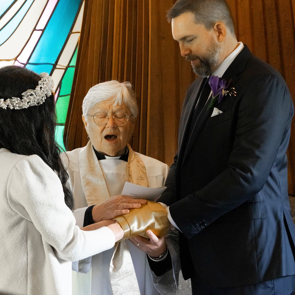 A wedding ceremony takes place in front of a stained glass window with an officiant joining hands.