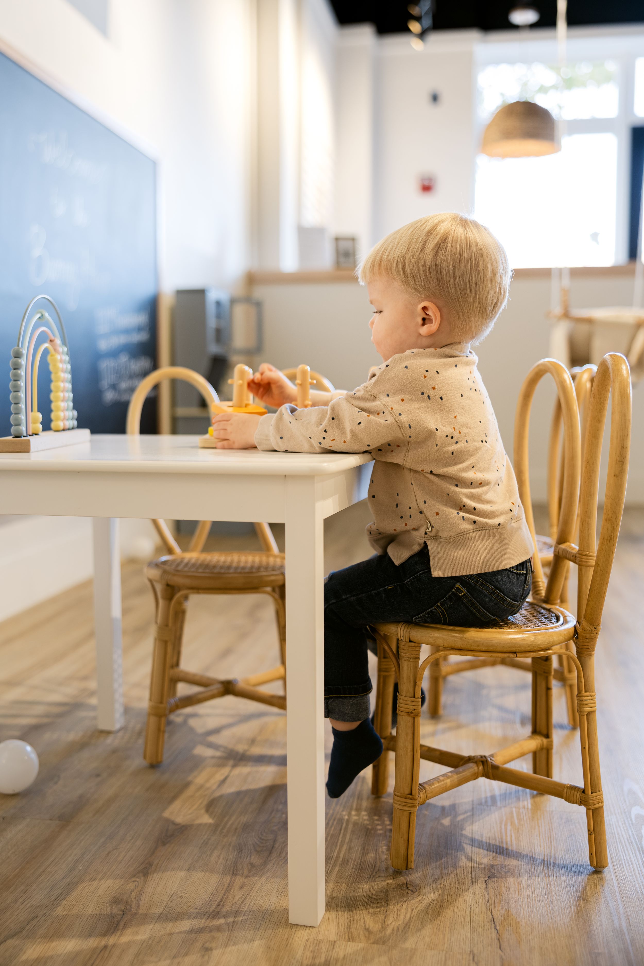 Small child sitting in a wooden chair at a white table on hardwood floors in a bright interior space.