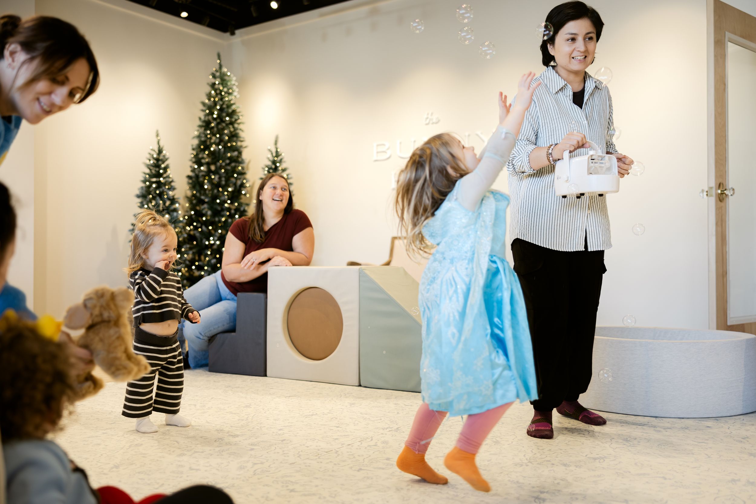 Children and adults participate in an interactive performance with Christmas trees in the background at an indoor venue.