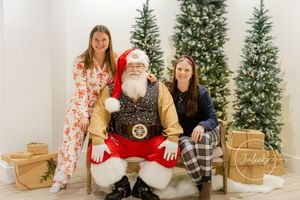 Holiday photo with Santa Claus seated between two people in festive attire with decorated Christmas trees in background.
