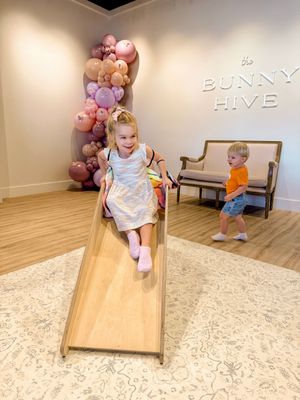 Two small children sit and play on a wooden slide in a room decorated with pink and purple balloons.