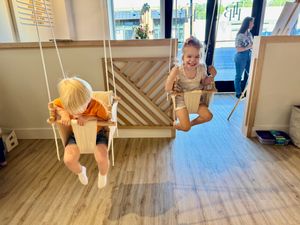 Two people swing on indoor swings mounted in a modern room with wooden floors and natural lighting.
