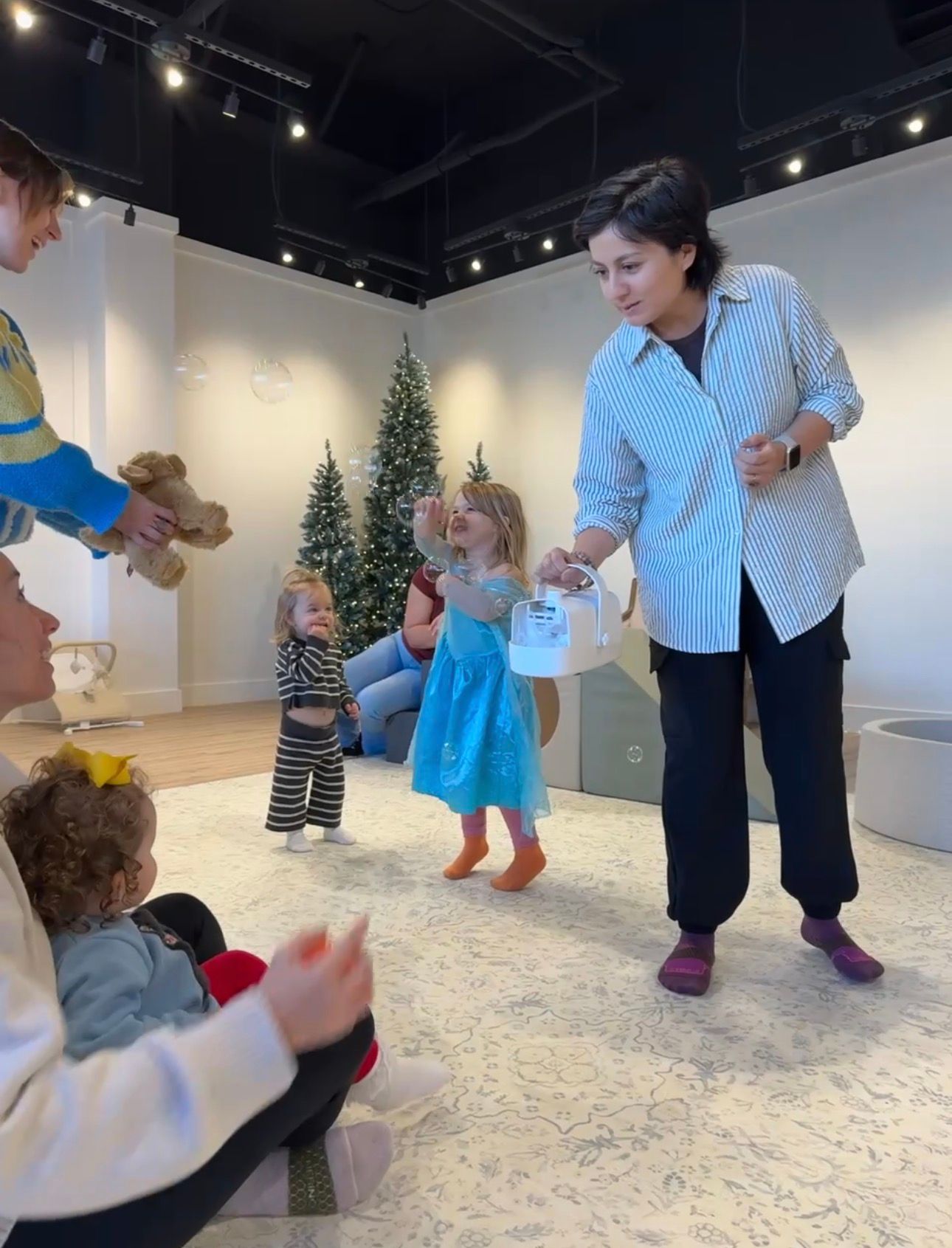 A group of children and adults participate in a dance class on a white floor in a studio with Christmas trees in the background.