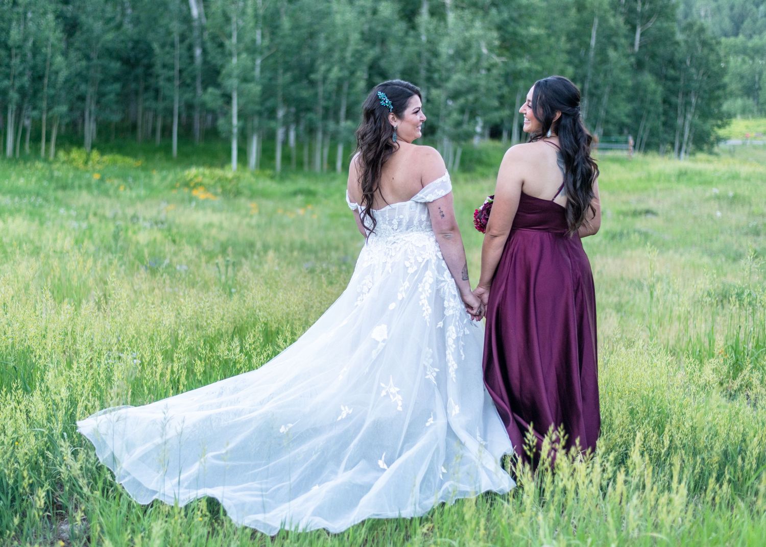 Two brides holding hands in a lush green meadow, one in white lace gown and one in burgundy dress.