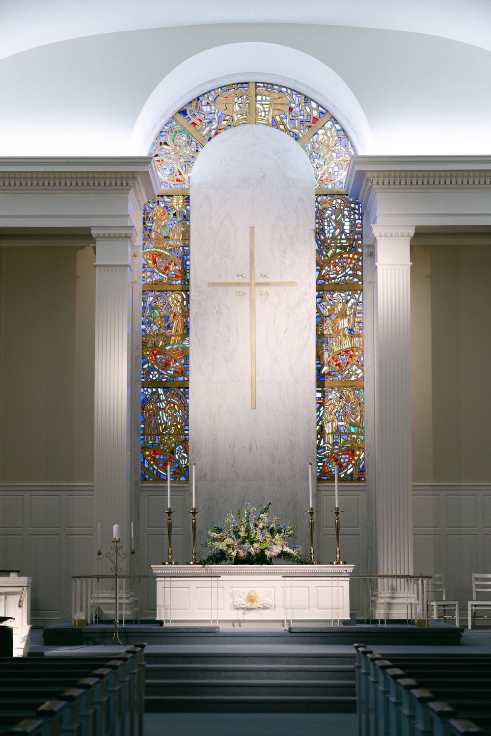 White church altar with stained glass windows, cross, and candles in an elegant chapel setting.
