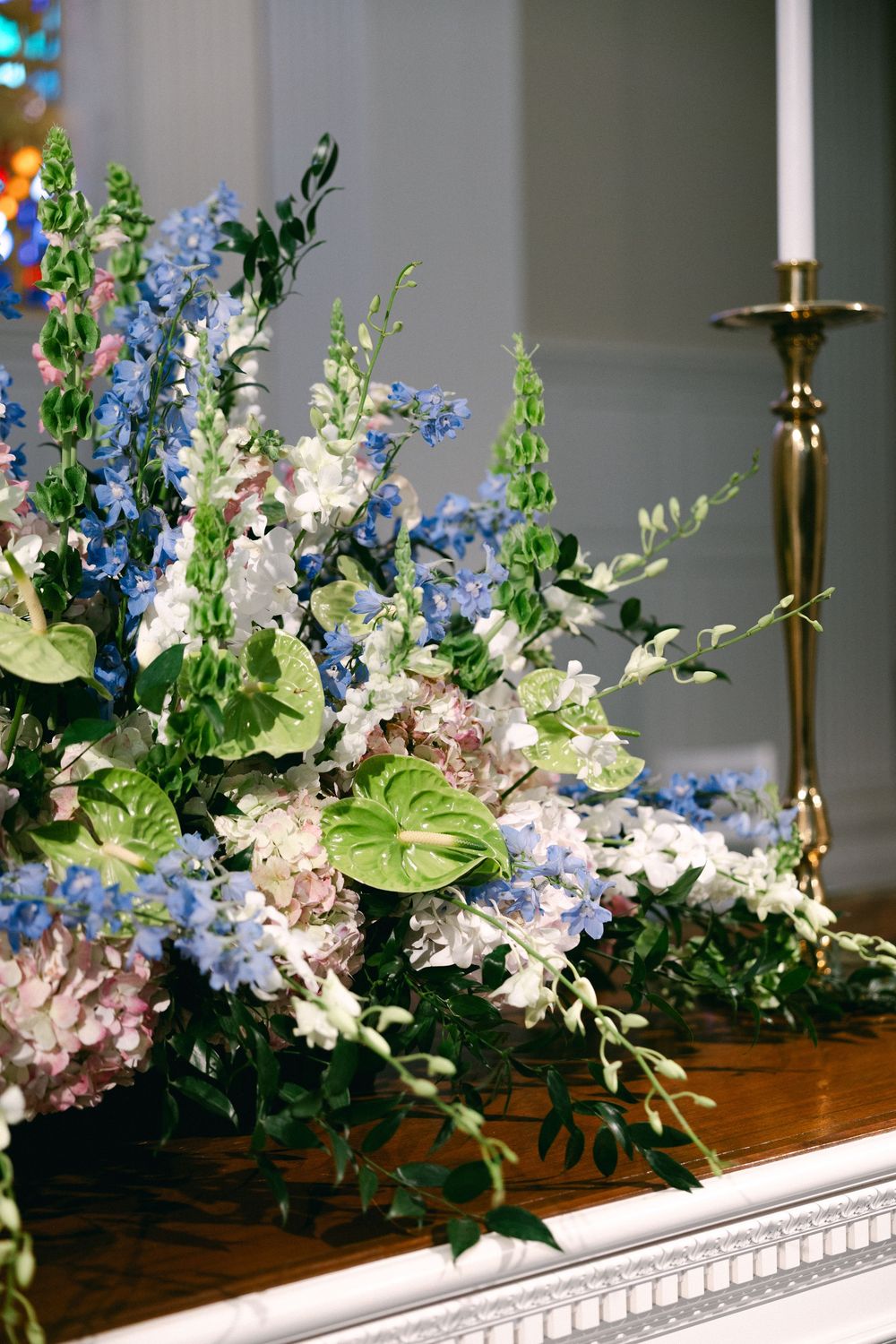 Elegant floral arrangement with blue, white and pink flowers displayed on a wooden surface with brass candlesticks.