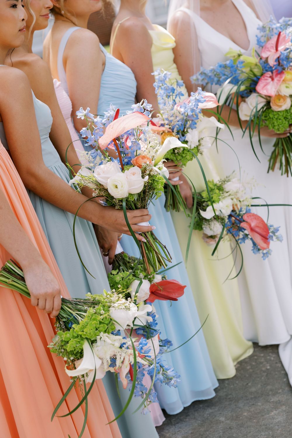 Bridesmaids in pastel dresses of coral, blue and yellow holding elegant floral bouquets at a wedding ceremony.