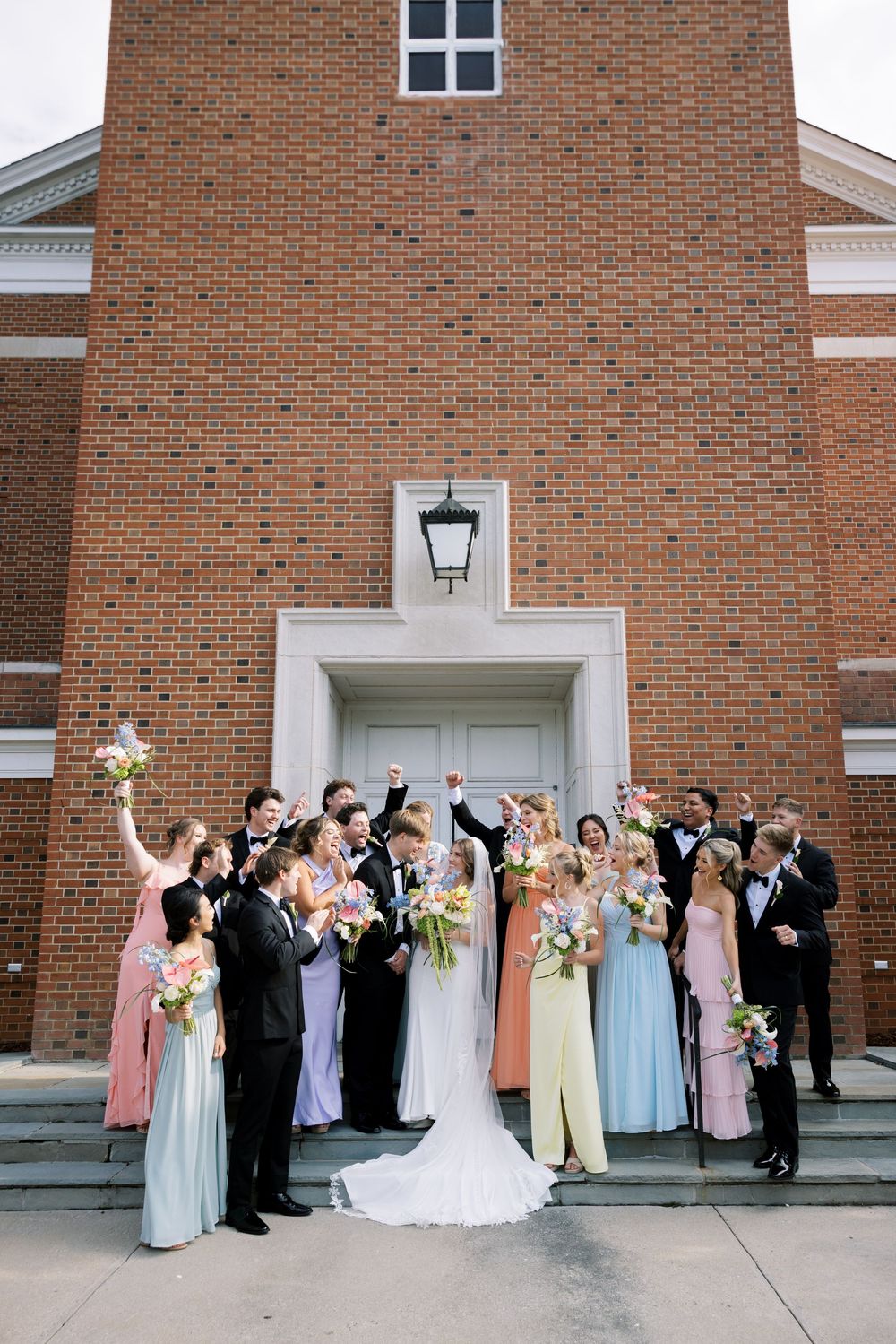 Wedding party poses outside brick church building in pastel bridesmaid dresses and formal attire.