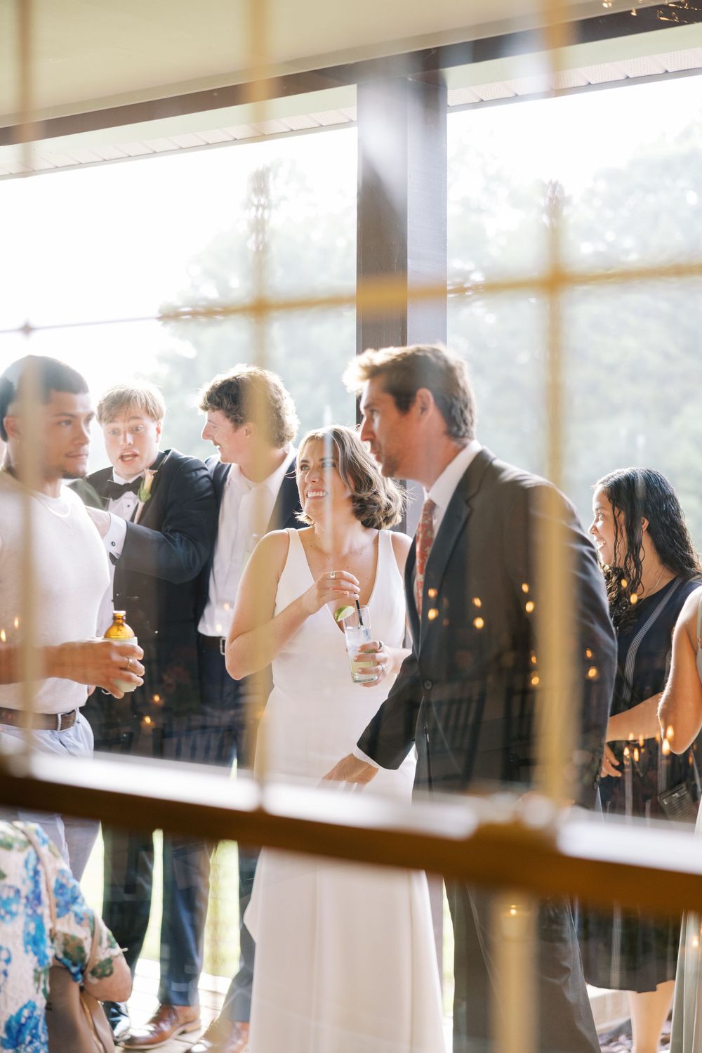 Wedding guests gather near sunlit windows for drinks and conversation at an elegant reception.