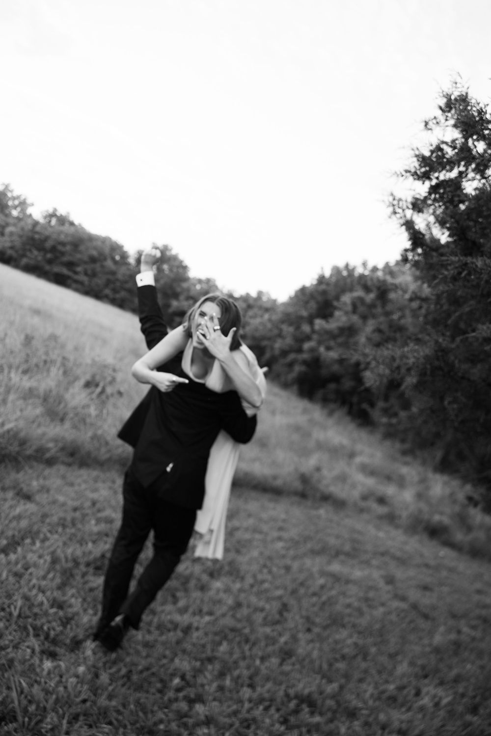 A black and white photo of a couple dancing in a grassy field with trees in the background.