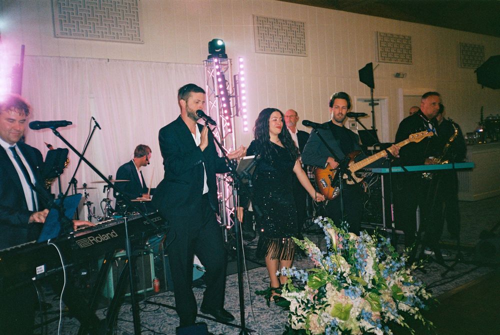 Live band performs on stage with instruments and microphones next to floral arrangements at an indoor event.