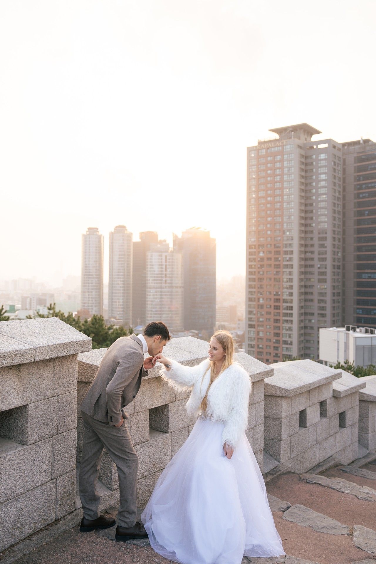 A couple in wedding attire poses for romantic photos on a rooftop with city buildings in the background at sunset.