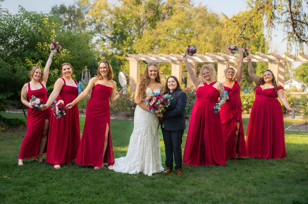 A wedding party in red bridesmaid dresses and white bridal gown poses outdoors in a garden setting during autumn. Quad Cities Wedding Photog