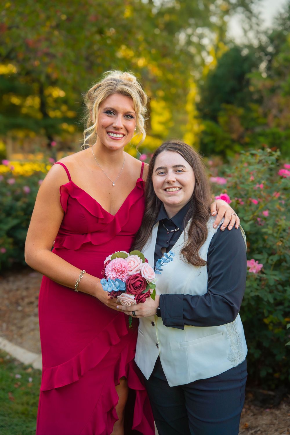 Wedding party members in red dresses and formal attire pose together in a garden with colorful fall foliage.