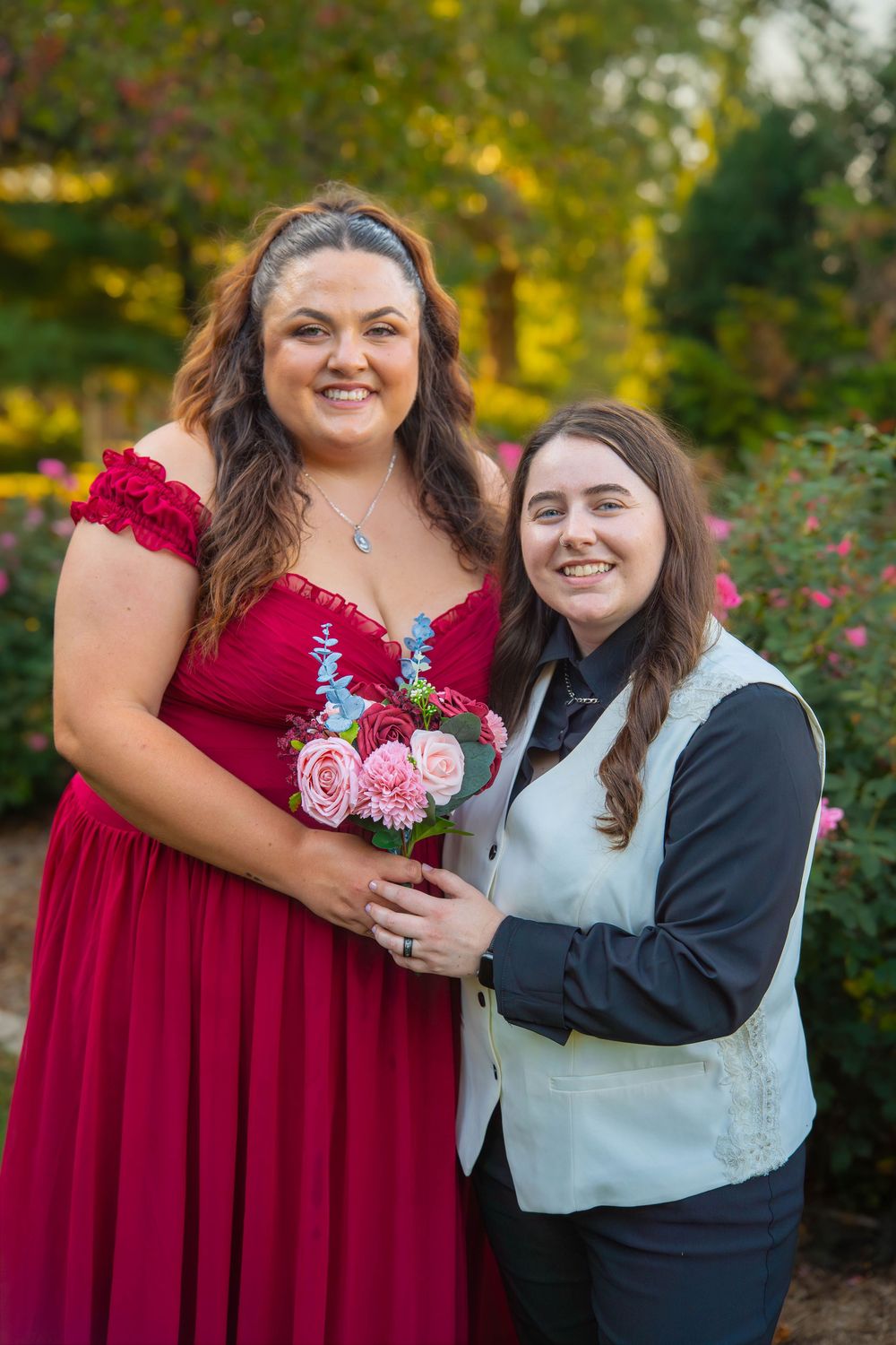A couple poses together in a garden, one wearing a red formal dress holding pink roses while the other wears a vest.