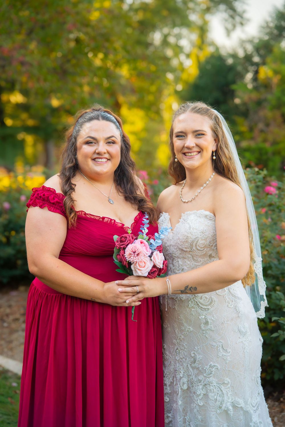 Two people in red and white formal dresses pose together in a garden setting with autumn foliage.