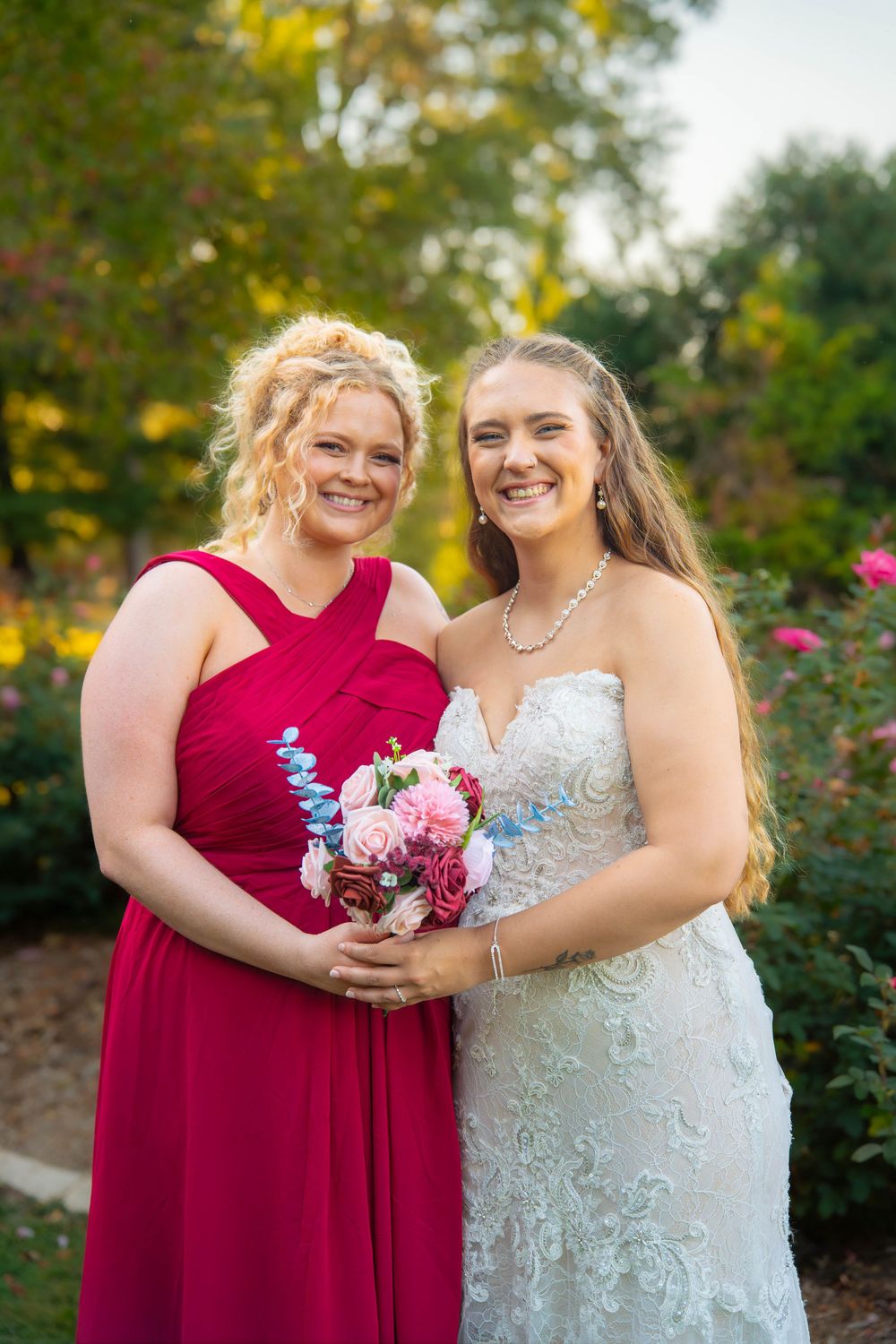 Two people in formal attire, one in a red dress and one in white, pose together in a garden with autumn foliage.