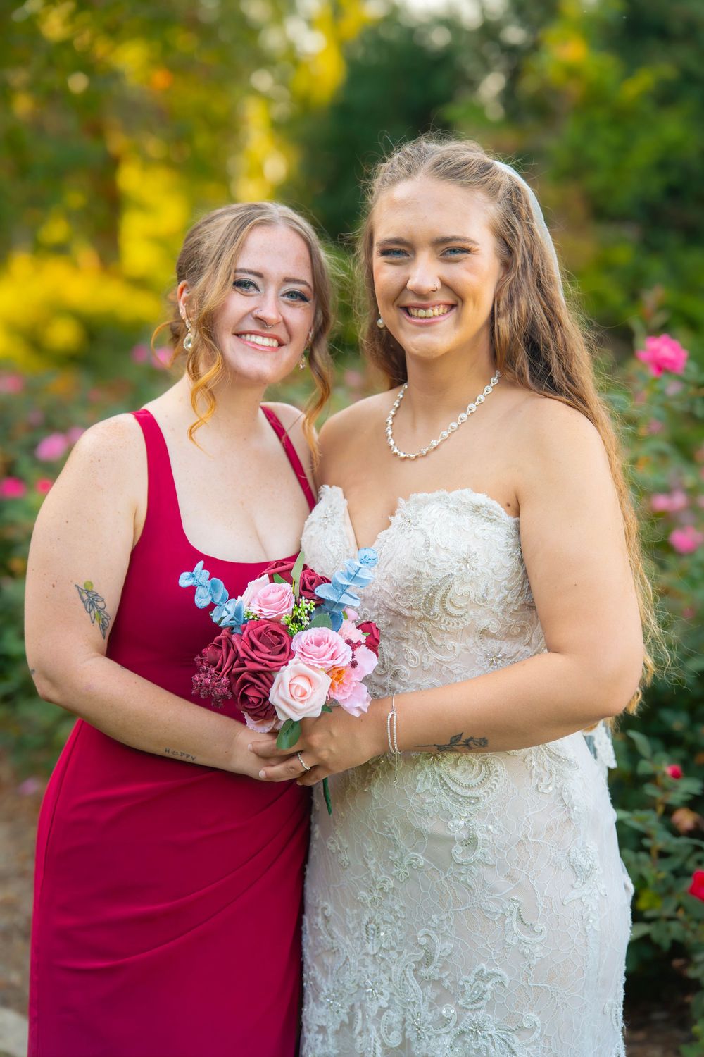 Two women pose together at a wedding, one in a red bridesmaid dress holding flowers and one in a white lace gown.