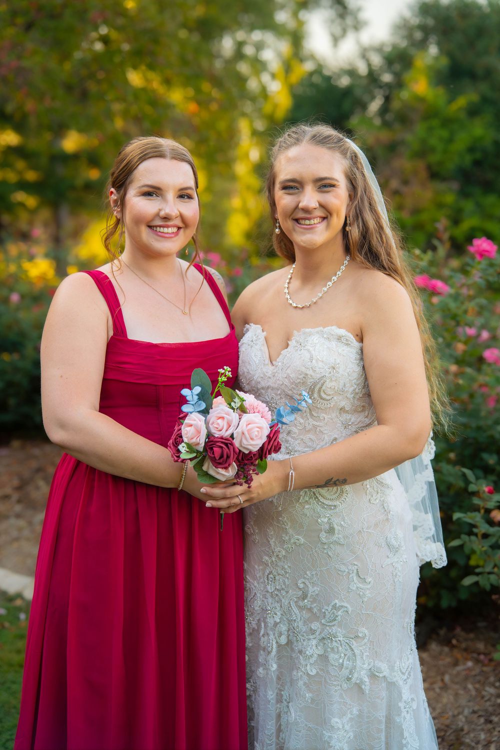 Two people in red and white dresses smile while holding bouquets in an outdoor garden setting.