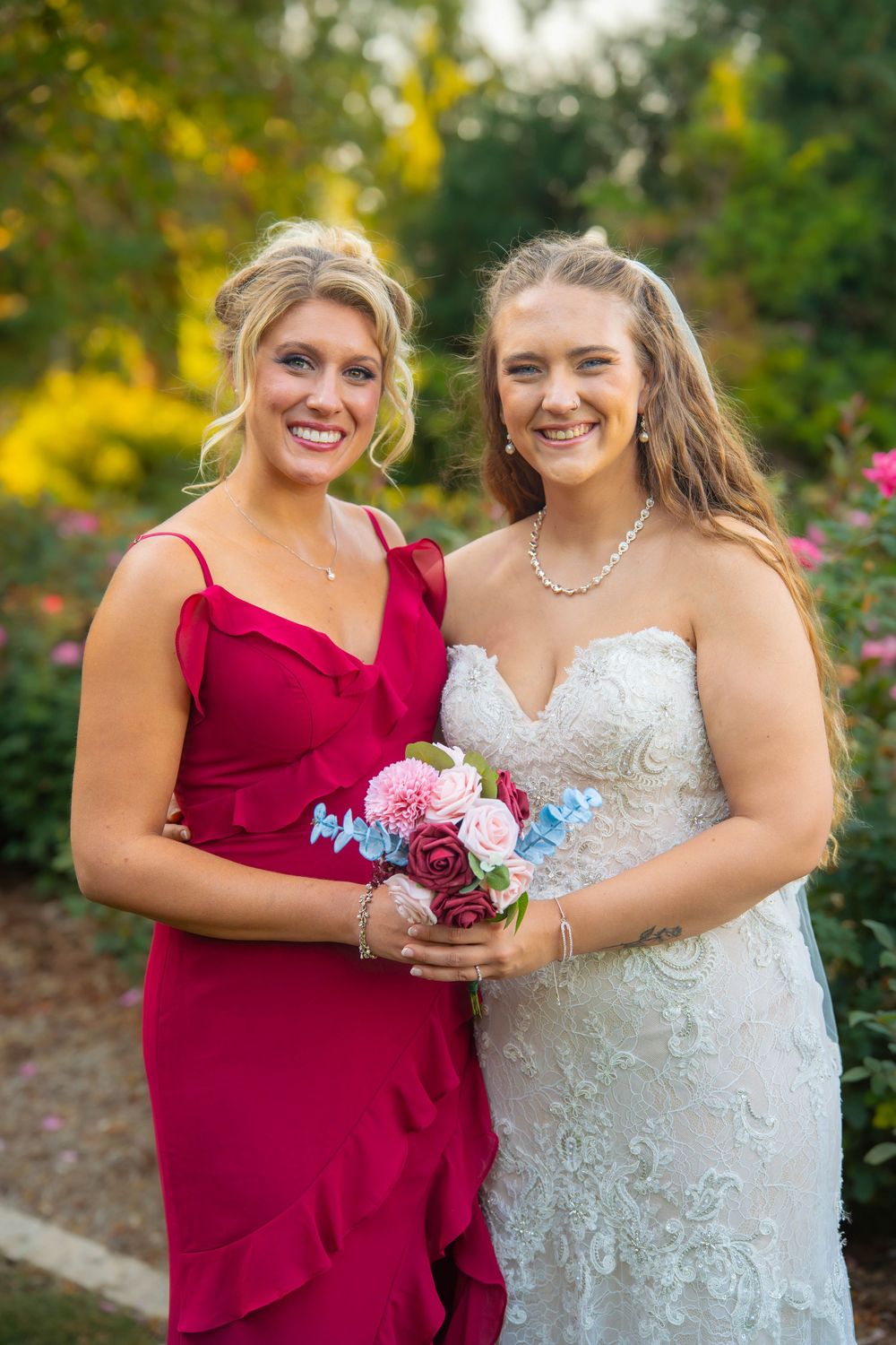 Bridesmaids share joyful moments while holding pink and burgundy floral bouquets in an outdoor garden.