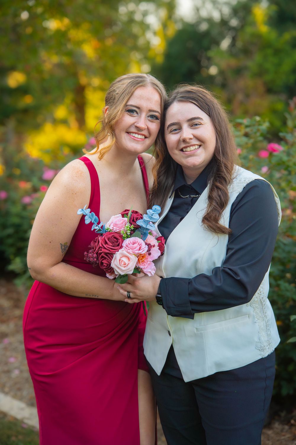 Two people in formal attire share a joyful moment outdoors with pink floral bouquets against a garden backdrop.
