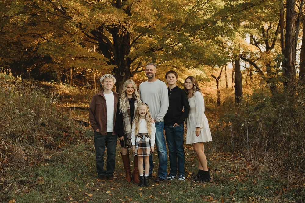 A family poses together in a wooded area during autumn with golden sunlight filtering through the trees.