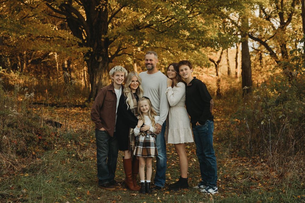 A group of people stand together in a wooded area during golden hour with autumn foliage in the background.