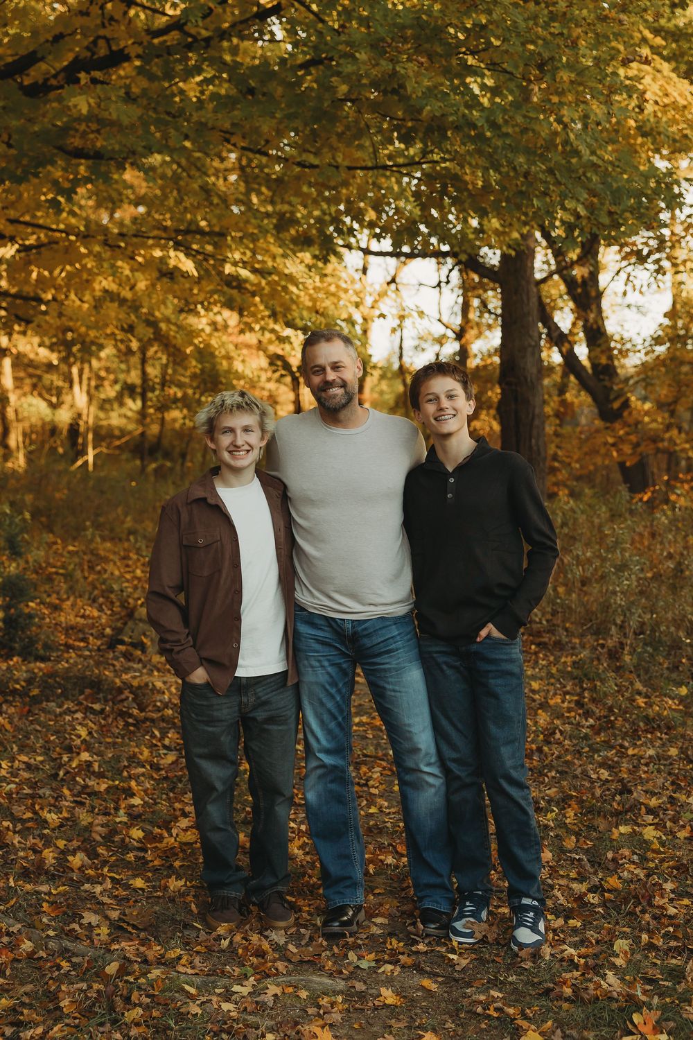 A warm autumn portrait of three people standing together on fallen leaves under golden trees in a park.