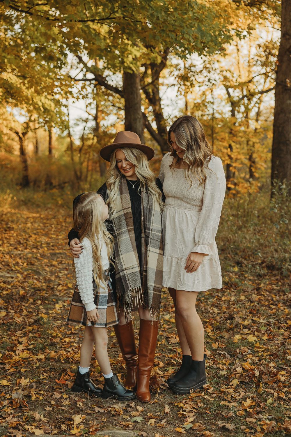 Couple in autumn forest wearing cozy sweaters and boots stand among fallen leaves at golden hour.