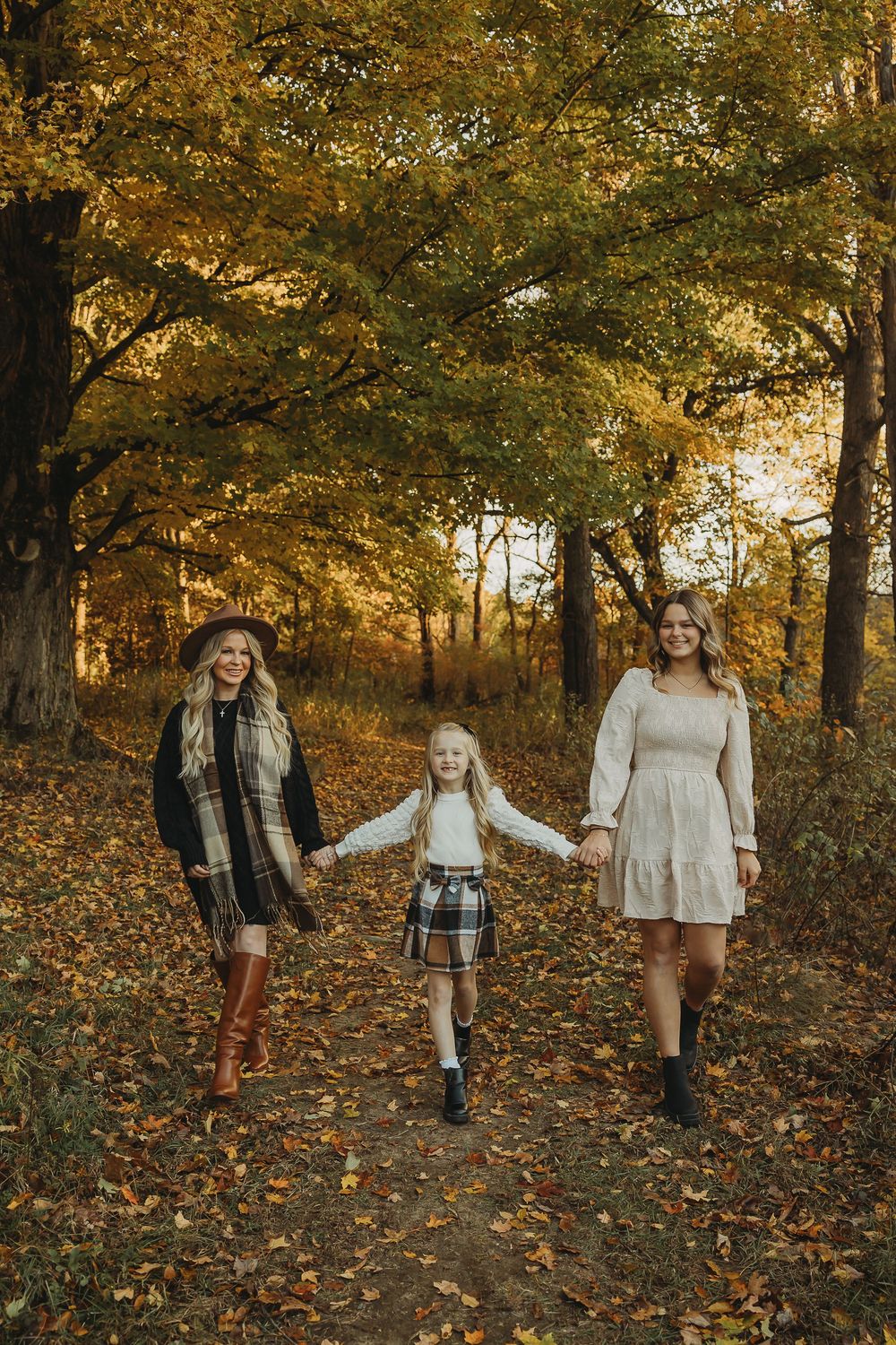 Three figures walking hand in hand down a leaf-covered forest path on an autumn day with golden foliage overhead.