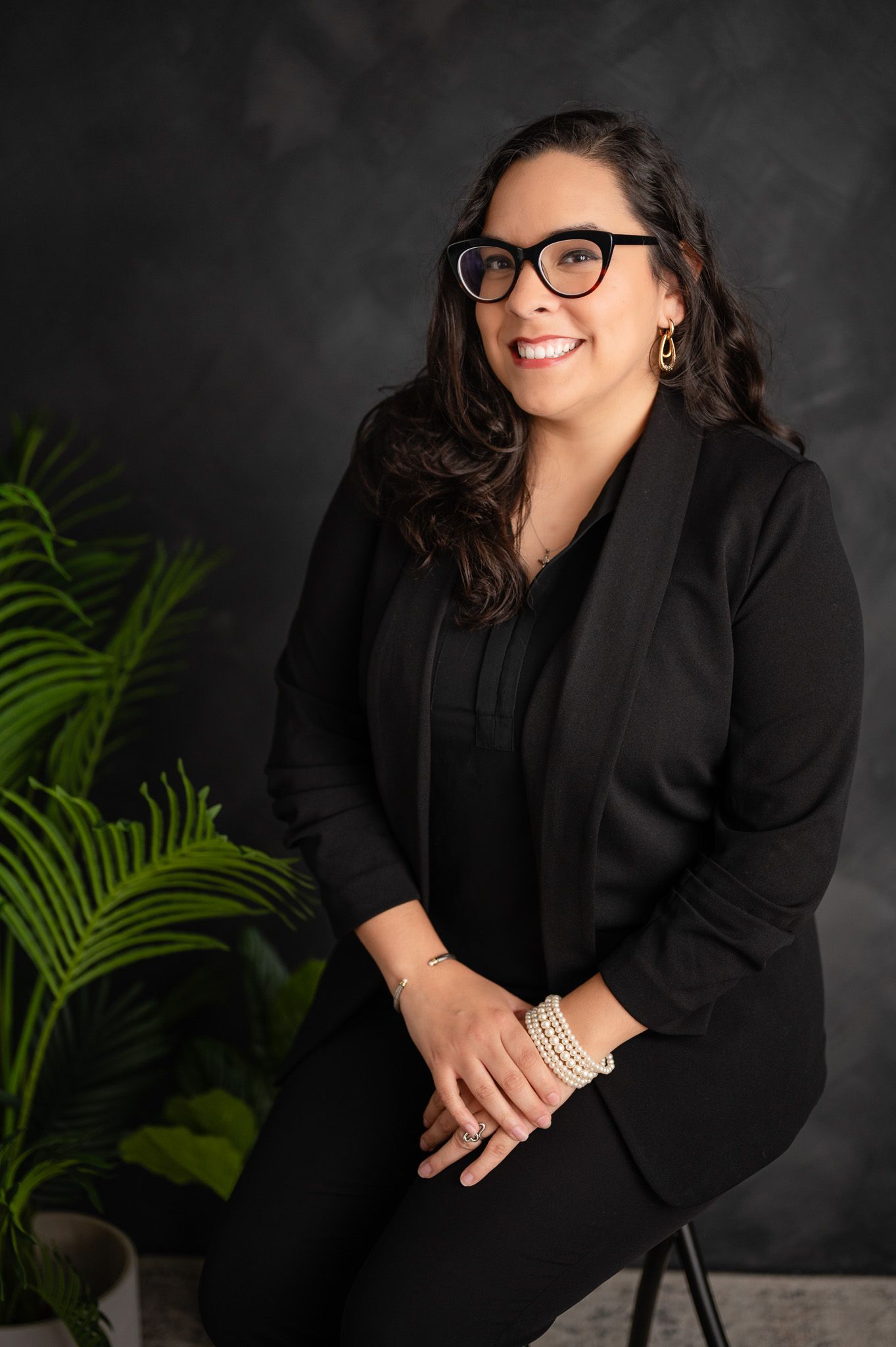 Professional studio portrait of a woman in black business attire and glasses seated with palm plants against dark backdrop.