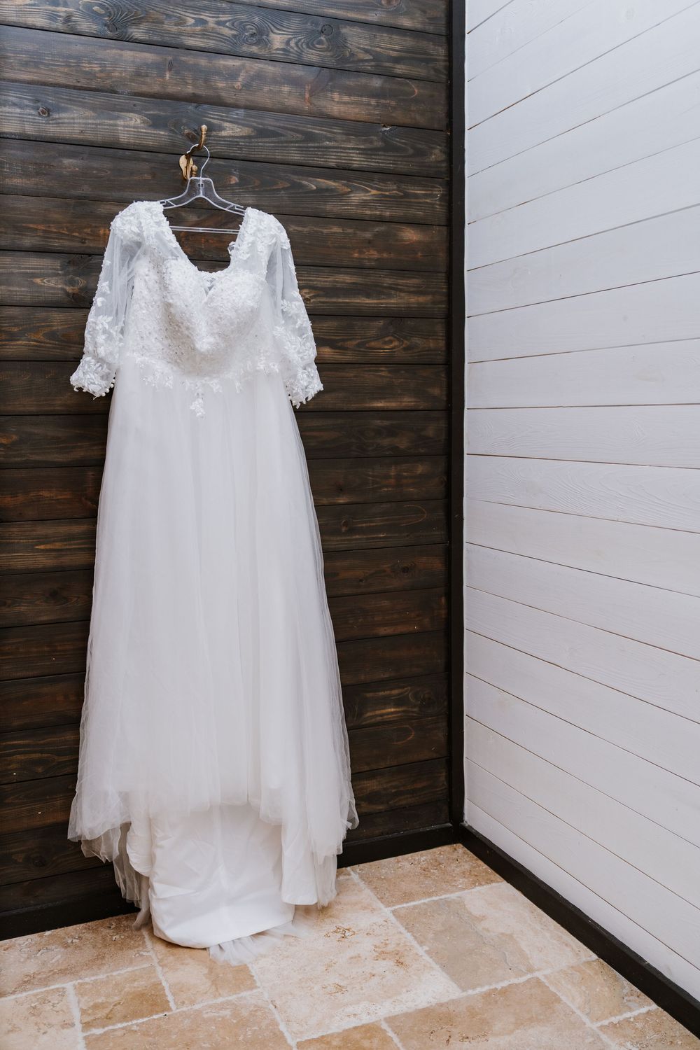 A delicate white lace wedding dress with sheer sleeves hangs on display against a rustic wooden wall.