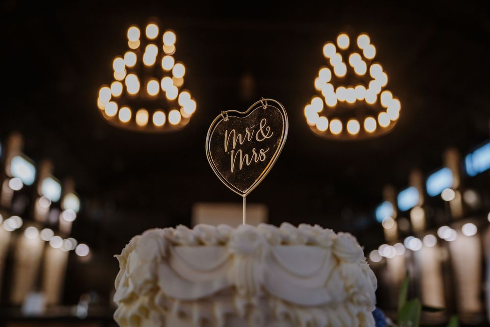 Wedding cake display with heart-shaped topper and string lights in dark romantic setting.