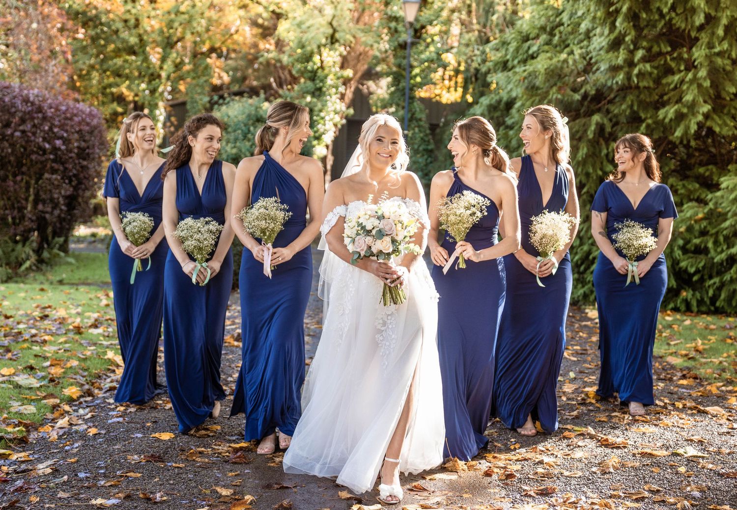 Wedding party in navy blue dresses and white gown poses together on autumn path covered with fallen leaves.