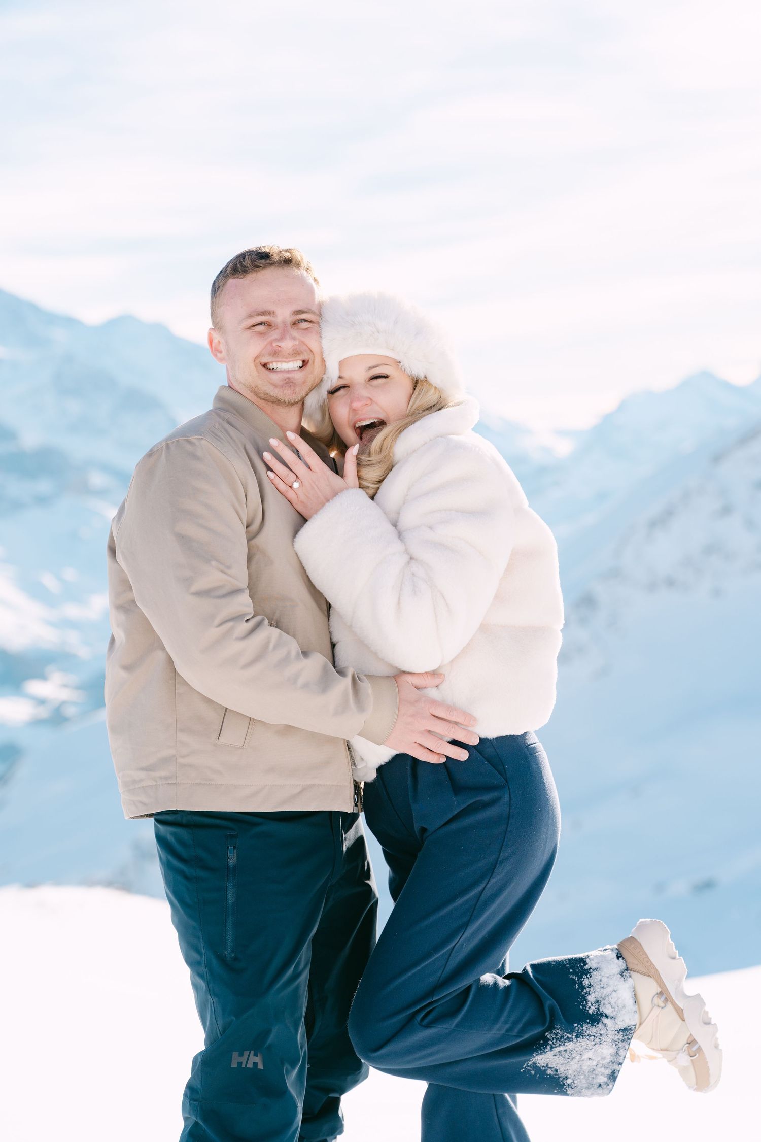 A couple shares a romantic moment in the snowy mountains wearing matching beige and navy winter outfits.