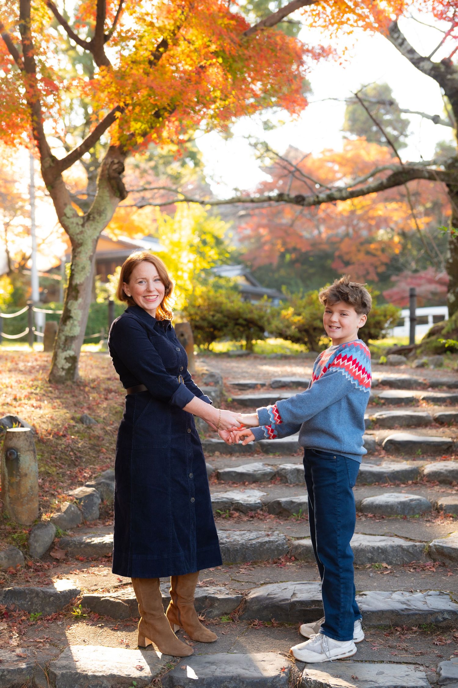 A mother and son hold hands and smile on stone steps amid stunning fall foliage.