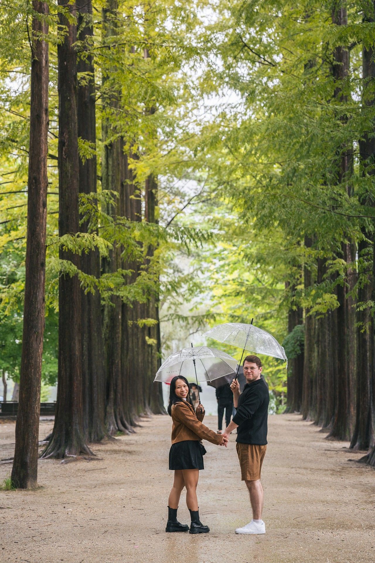 Two people holding umbrellas stand on a path lined with tall green trees in a romantic autumn setting.