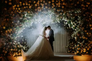 Dreamy wedding scene under floral arch with dramatic lighting and bokeh effects.
