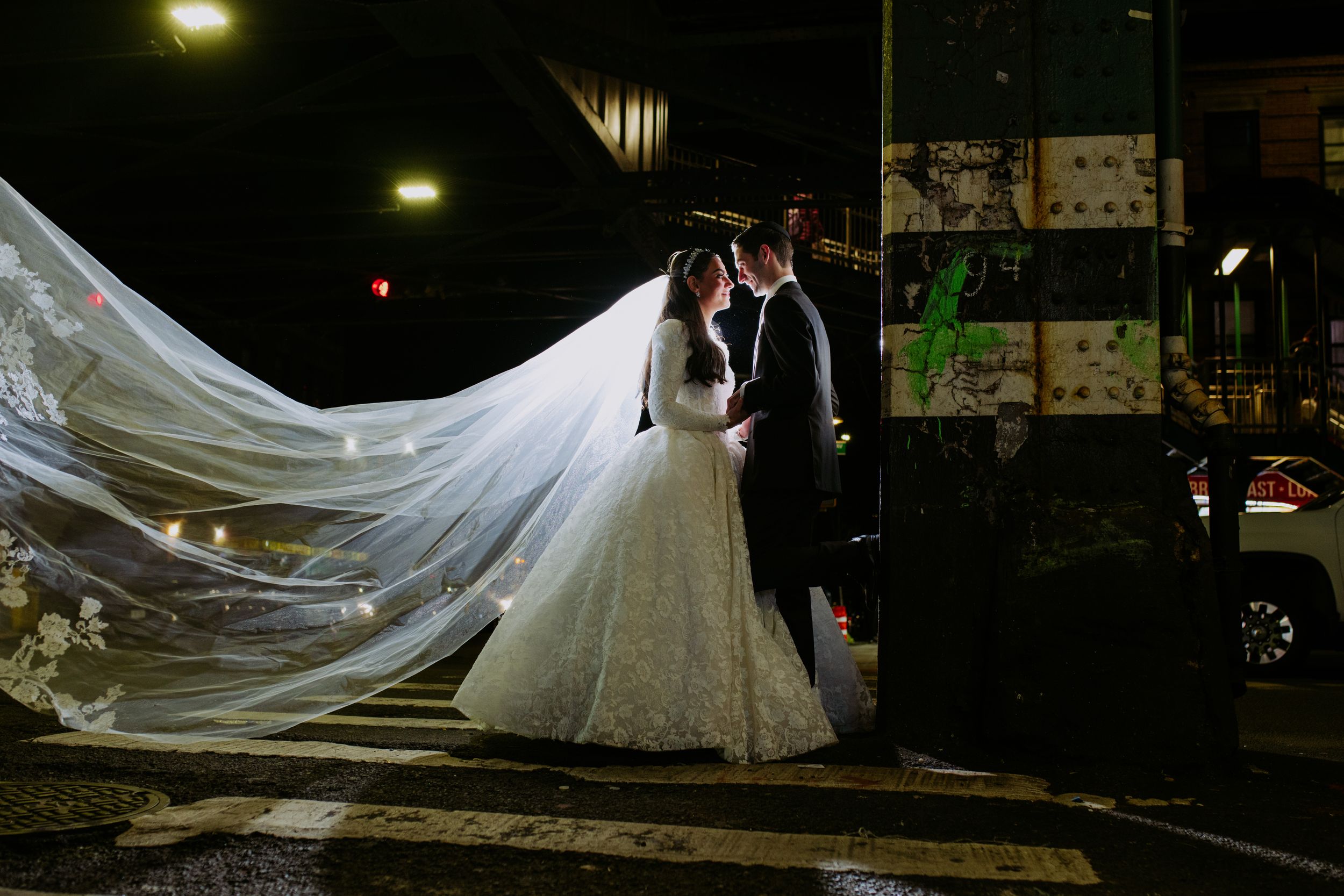 A bride and groom share an intimate moment under dramatic lighting while her long veil flows in the evening breeze.